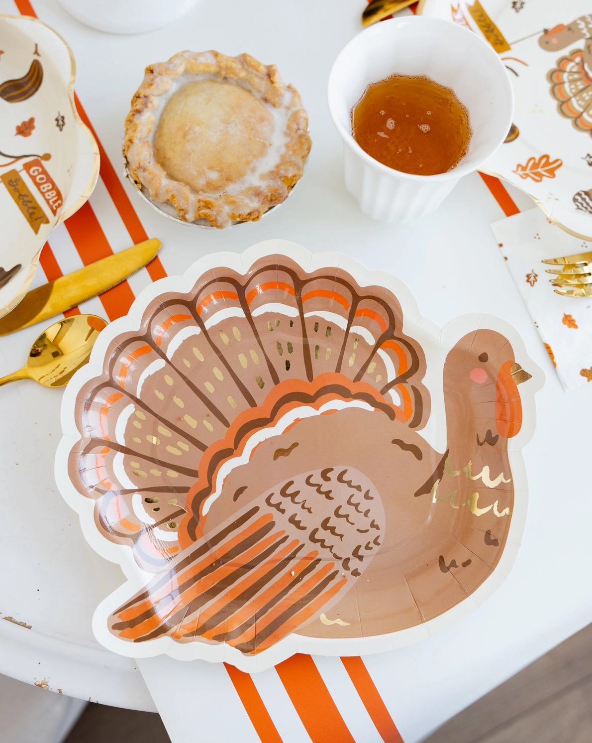 Turkey-themed paper plate on a table with a pie and cup of sauce.