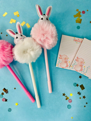 A set of Easter party favors including pink and white tutu bunny pens and bunny-themed notepads, displayed on a blue background with confetti.