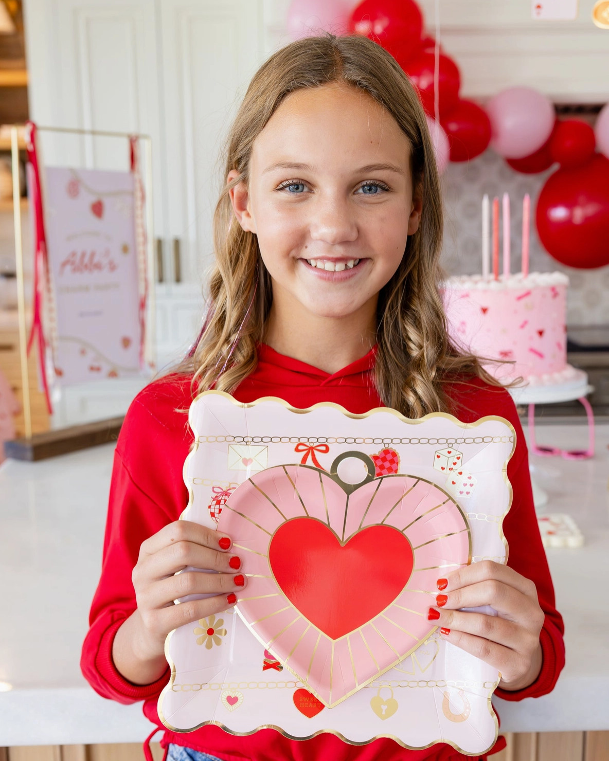 Young girl holding a heart-shaped cookie in a festive setting with balloons and a cake.