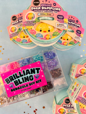 Toy glitter kit products arranged on a light blue background with star confetti. Visible items include multiple pink and mint packages labeled Hello Duck Dumpling Mystery Spushy Toy, a clear plastic case filled with assorted glitter colors labeled Brilliant Bling Bedazzle DIY Kit, and additional packaged kits partially visible at the edges.