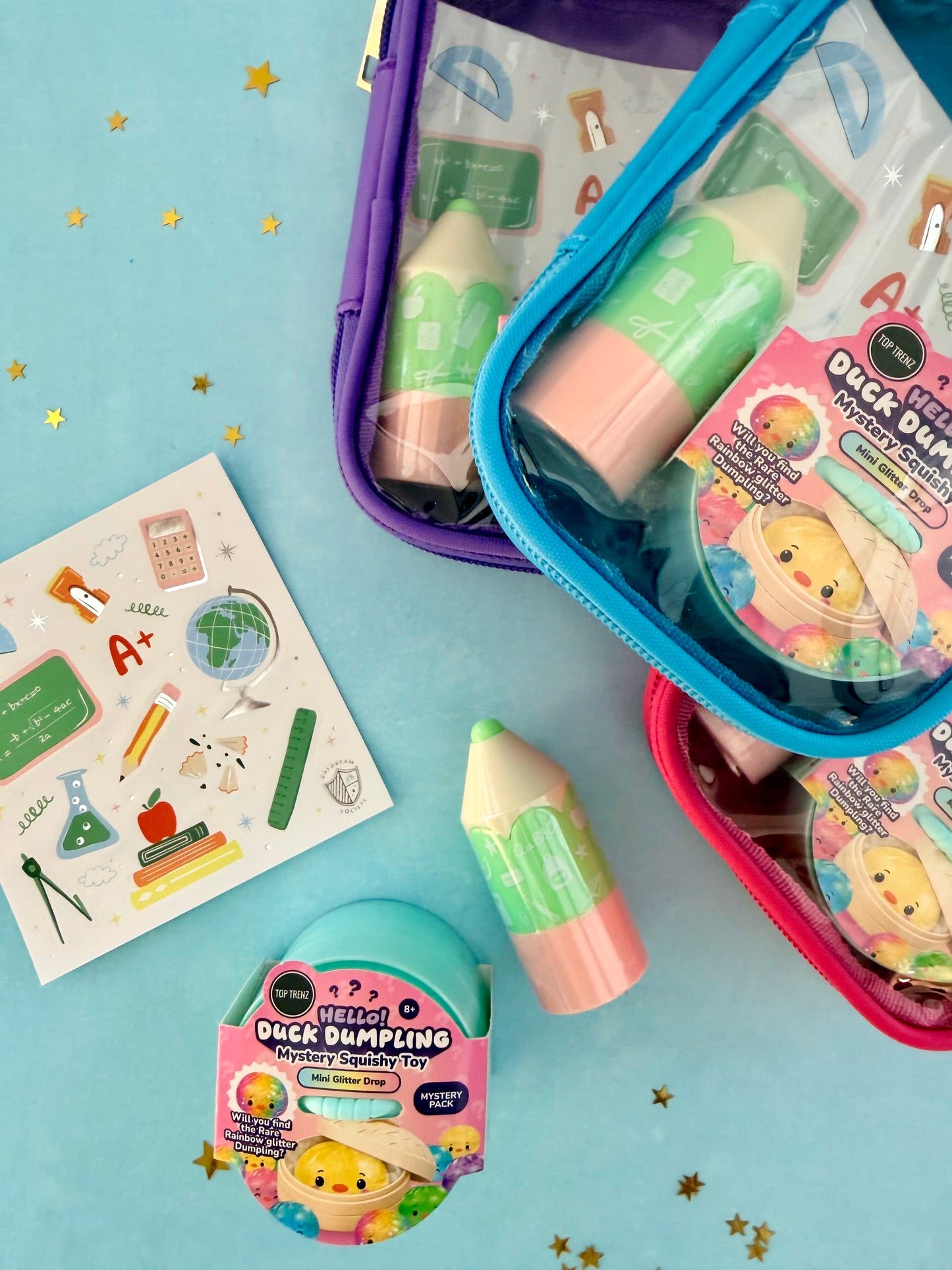 Three colorful zippered mystery pouches containing school-themed novelty items, arranged on a light blue background with gold star confetti. A sheet of educational stickers and a rolled mystery bubble toy are also visible.