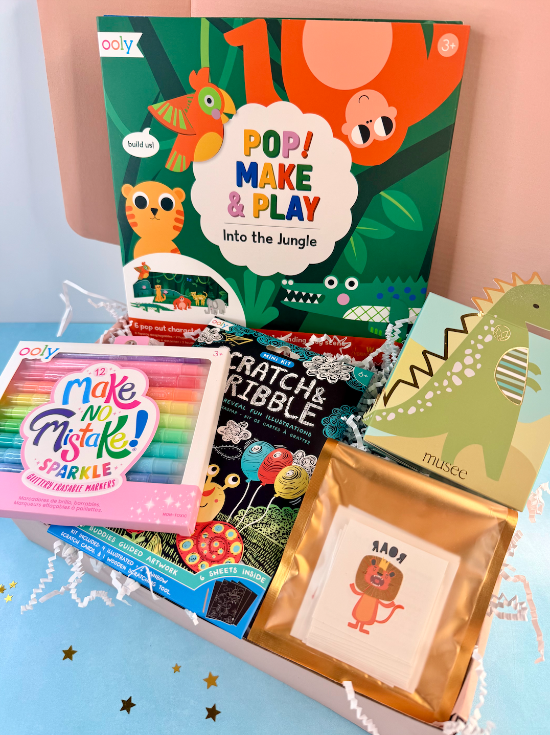 A curated children's gift box filled with colorful toys and books, including a large jungle-themed activity book titled 'POP! MAKE & PLAY Into the Jungle,' a pastel crayon set labeled 'make an Mistake! Sparkle,' a black-and-white picture book, a green dinosaur card, and a small gold-framed lion print.