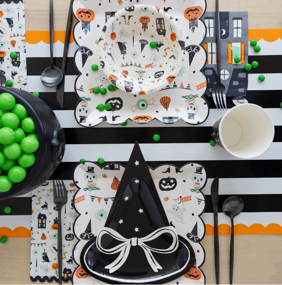 Halloween-themed table setting with plates, cups, and utensils on a black and white striped tablecloth.