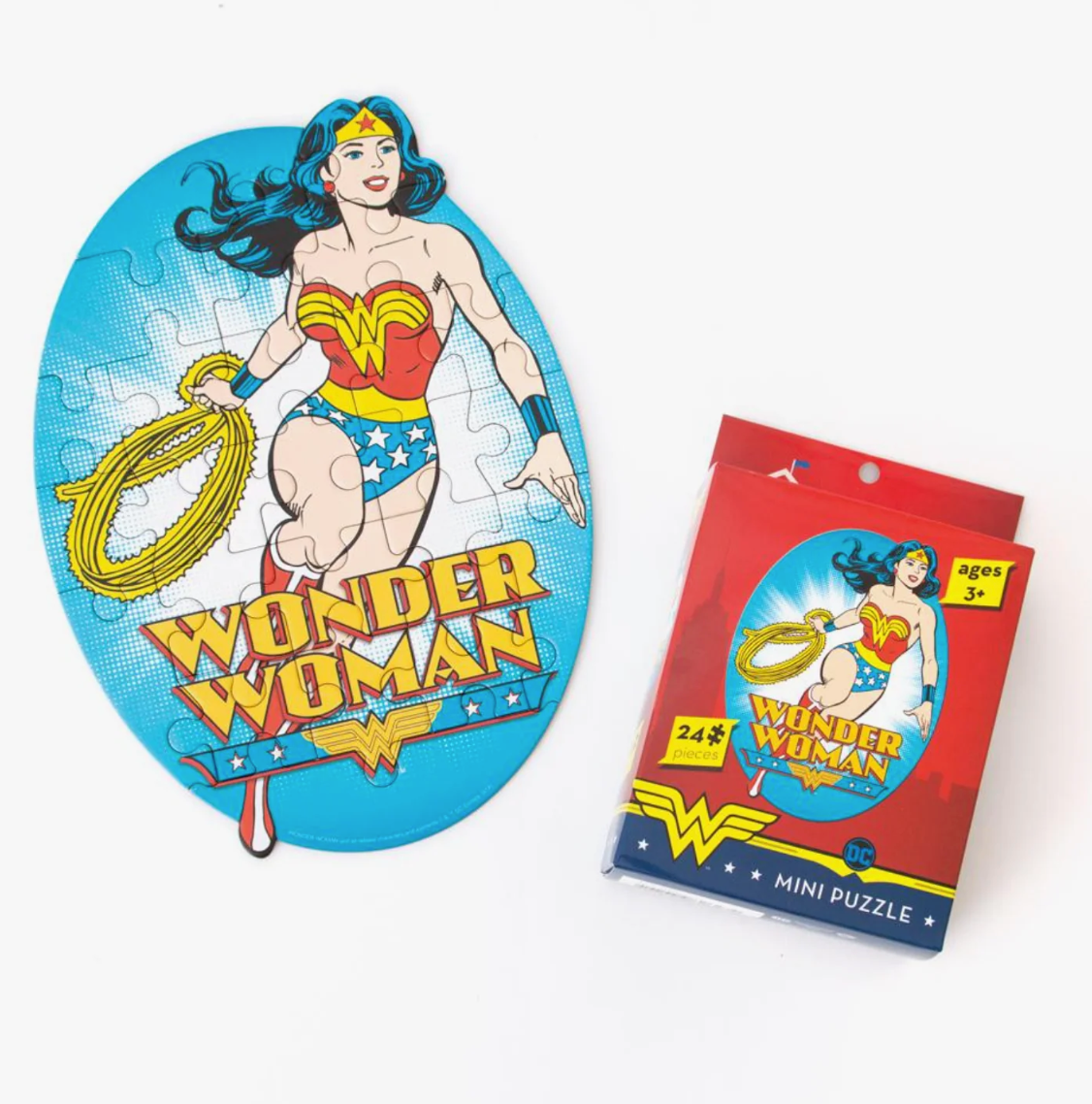 An oval jigsaw puzzle featuring Wonder Woman on a blue background with the words 'WONDER WOMAN' in large yellow letters, shown next to its red retail packaging on a white background.