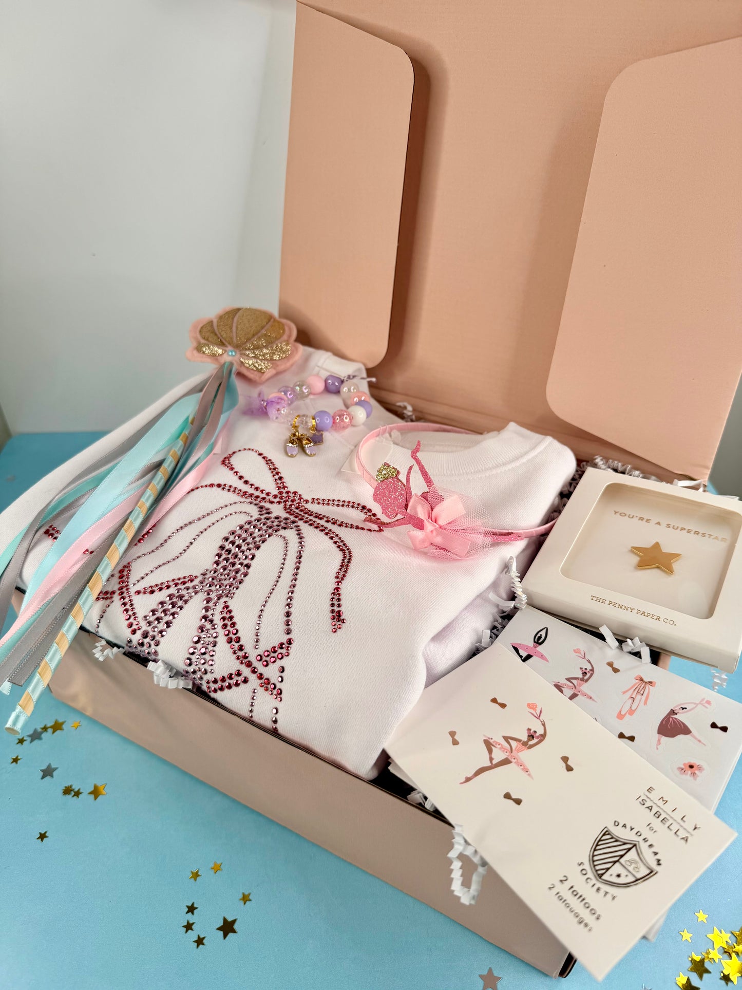 Open box containing a baby outfit with a bow, cards, and other items on a blue surface.