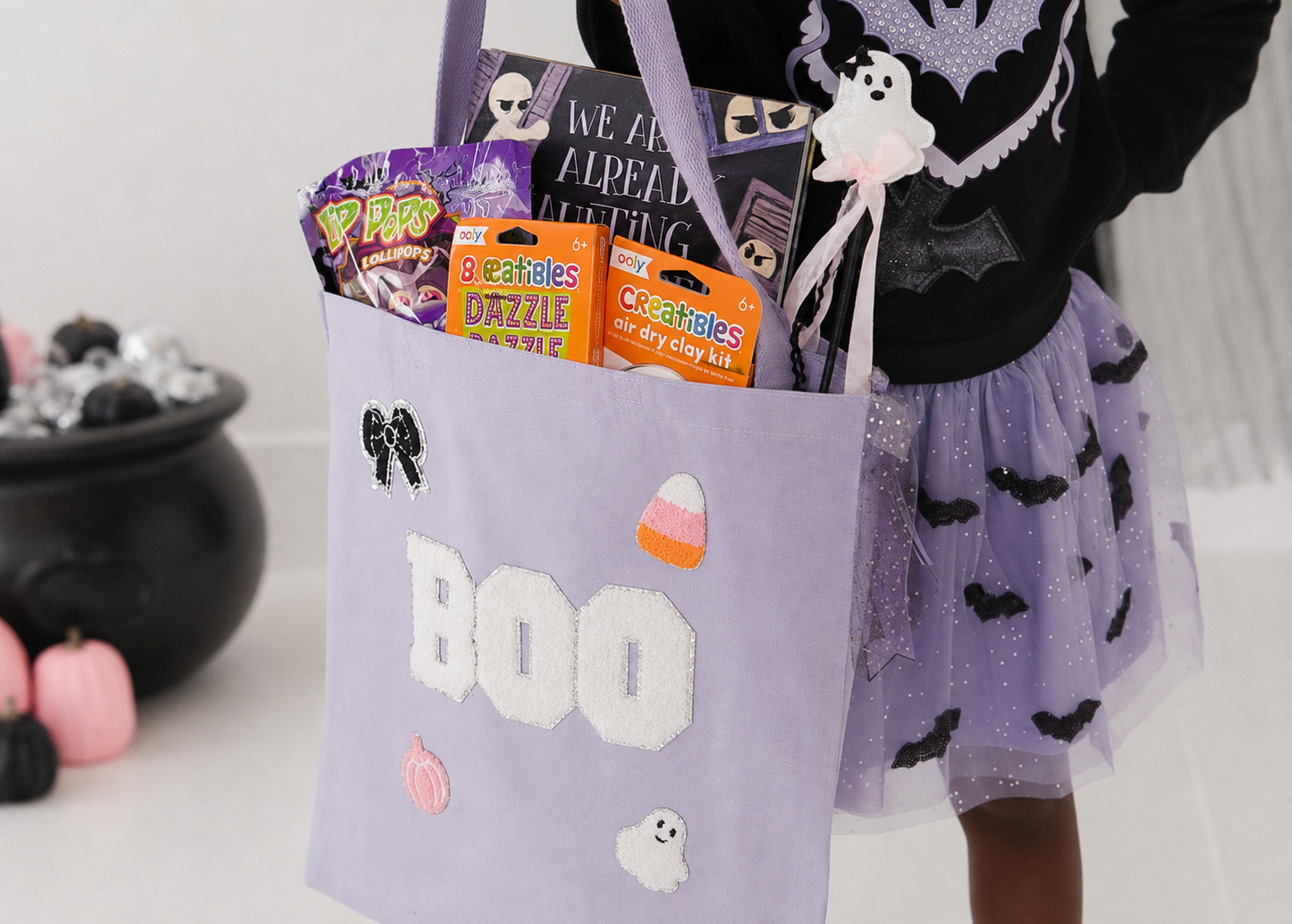 A child wearing a black and lavender Halloween outfit holds a lavender fabric trick-or-treat bag decorated with white 'BOO' lettering, black bat silhouettes, a ghost, and candy corn motifs. The bag is filled with treats, including a purple Pop-Ras and an orange Goochsticks kit box, against a white studio background with a black cauldron prop on the left.