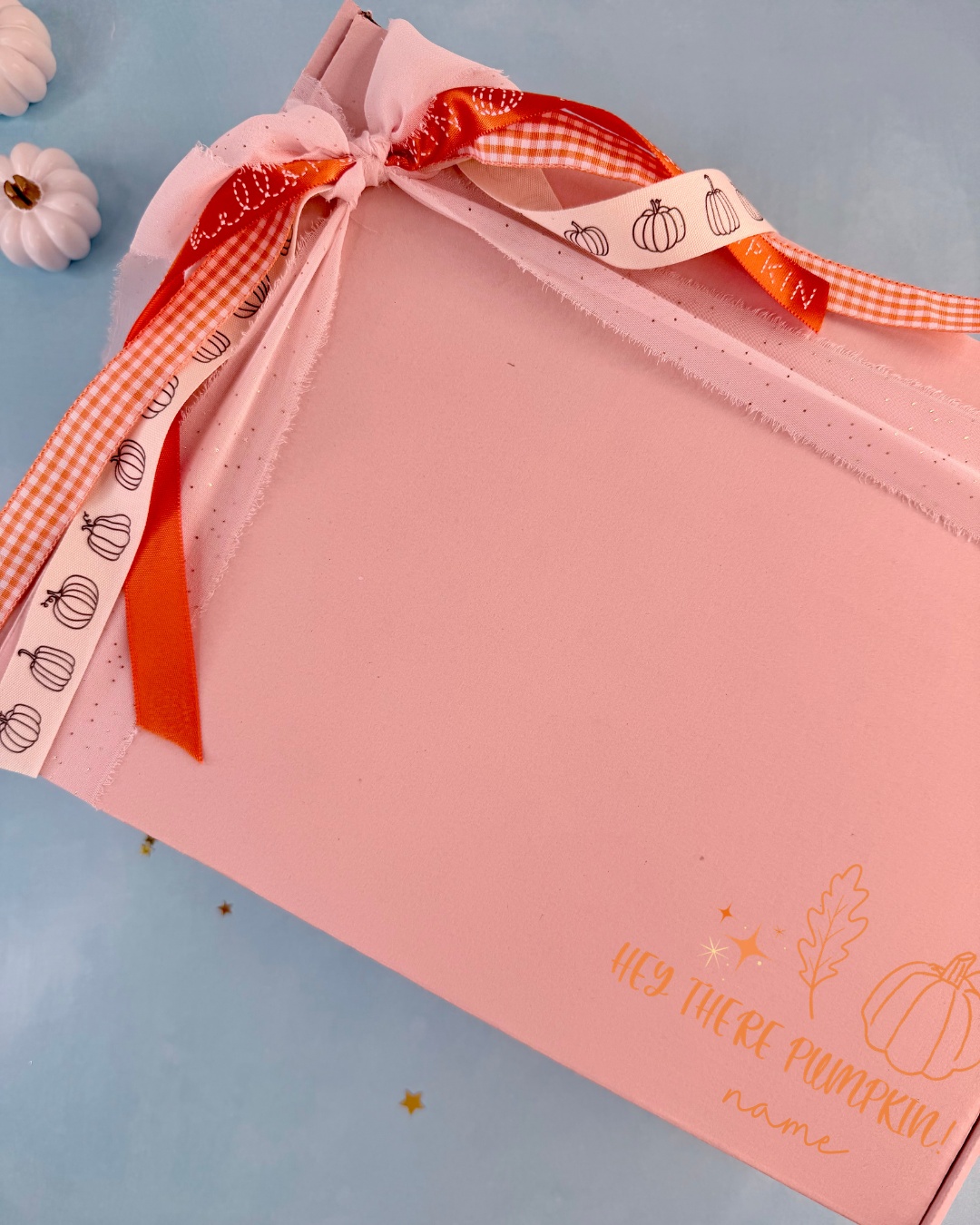 A pink gift box with a personalized name printed in gold on the front, tied with a decorative ribbon featuring orange gingham and white ribbon with pumpkin illustrations. The box is photographed on a light blue background with small decorative pumpkins in the corner.
