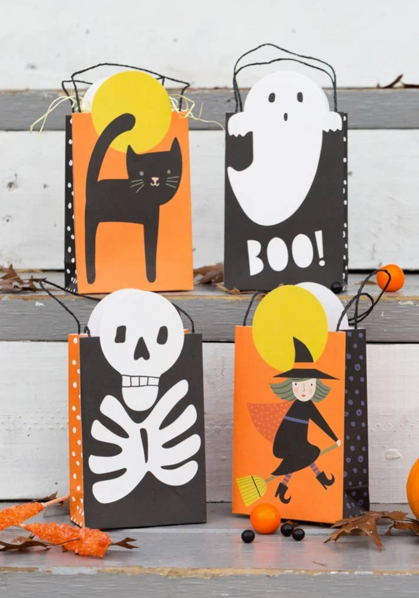 Four Halloween-themed paper treat bags arranged on rustic gray wooden steps. The bags are black and orange with white polka dots and feature illustrations of a black cat, a ghost with the word 'BOO!', a white skeleton, and a witch riding a broomstick.