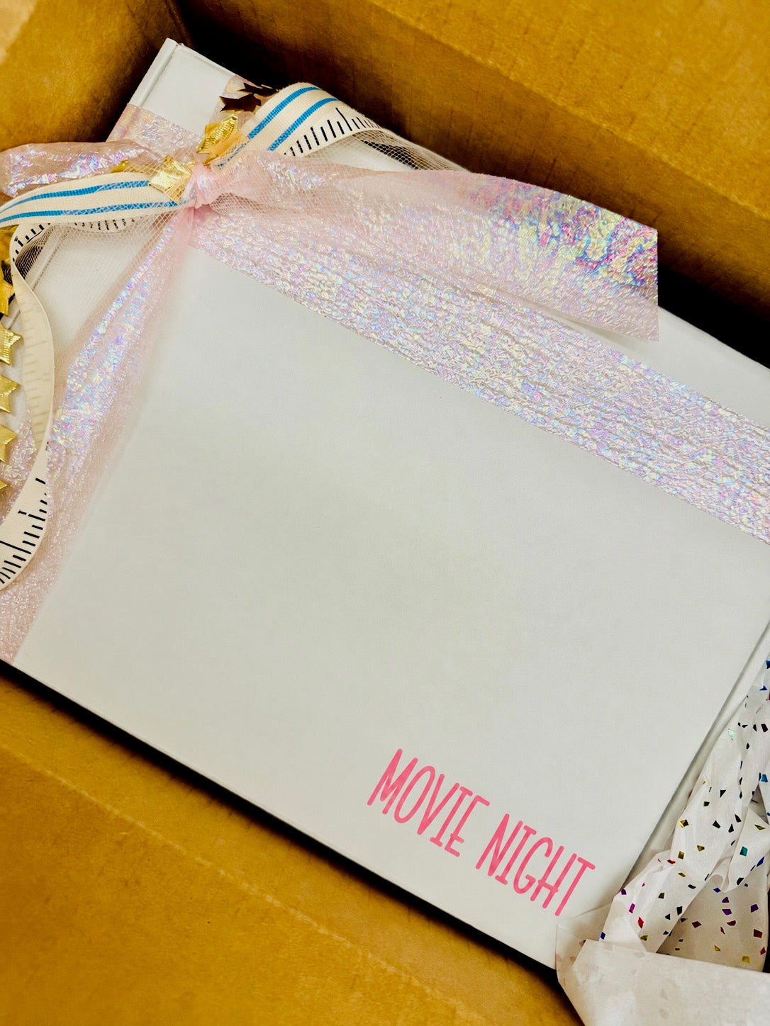 A white rectangular card or box insert with the words “MOVIE NIGHT” printed in pink at the bottom, tied with a sheer iridescent pink ribbon and placed inside a brown cardboard box. A small decorative gold charm is visible on the left edge.