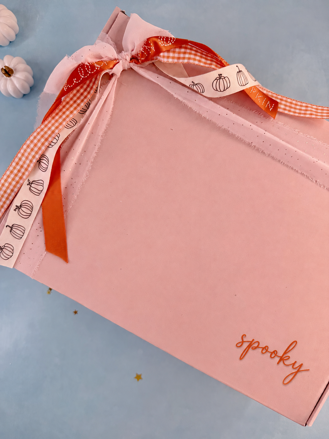 A pink rectangular gift box with the word 'spooky' printed in orange script near the bottom right, tied with a pink ribbon and orange gingham ribbon printed with pumpkin outlines, on a light blue background.