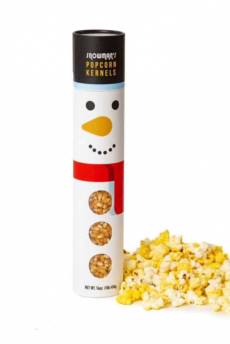 A snowman-themed popcorn kernel tube with a black top and a white body decorated with a smiling face, red scarf, and three circular windows showing the popcorn kernels inside. A small pile of yellow and white popcorn sits to the right on a clean white background.