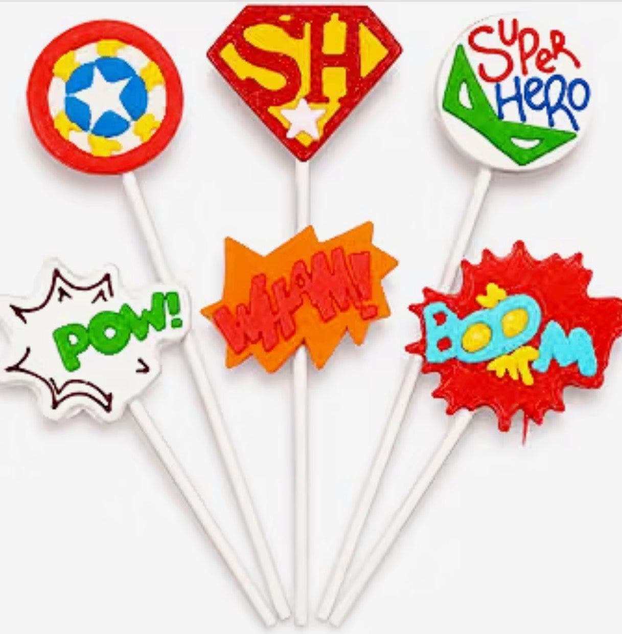 Six colorful superhero-themed lollipops on white sticks, featuring designs like a circular shield, a red 'SH' emblem, a green-and-blue 'Super Hero' sign, a white 'POW!' burst, an orange 'WHAM!' starburst, and a red 'BOOM!' explosion.