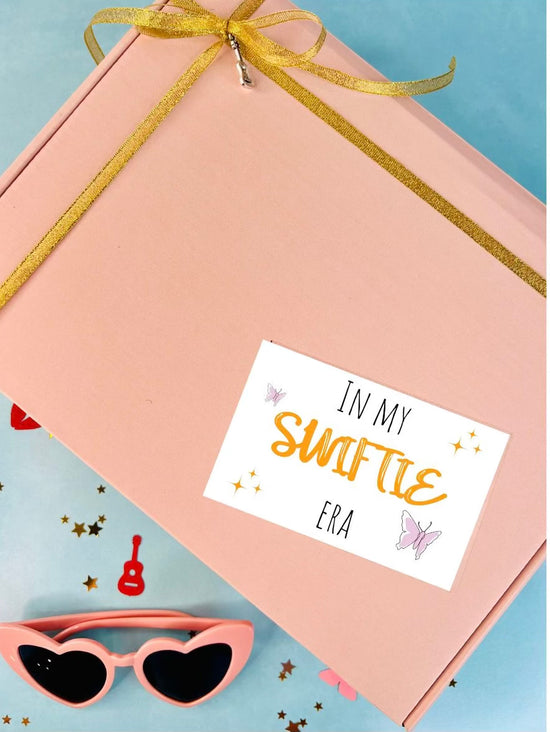 Pink gift box with gold ribbon, sunglasses, and 'In my Swifte Era' card on a blue background
