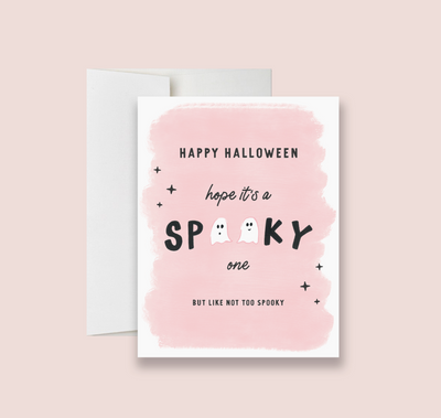 A folded Halloween greeting card with a pink background, text 'Happy Halloween' and 'I hope it's a spooky one BUT NOT TOO SPOOKY', and decorative elements like stars and a ghost. The card also includes a white envelope.